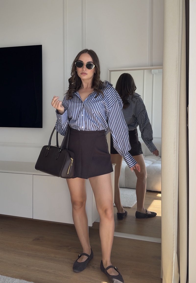 Femme portant des lunettes de soleil, un chemisier rayé, une jupe noire et des ballerines, tenant un sac à main noir, debout à l'intérieur près d'un miroir en pied.