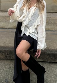 Blouse blanche à volants associée à une jupe asymétrique noire et des bottes noires hauteur genou. La tenue combine des éléments fluides et structurés.