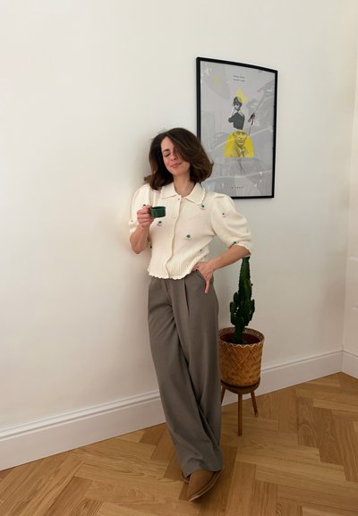 Mujer con suéter blanco y pantalones grises sosteniendo una taza verde, de pie cerca de un cactus en maceta y un cuadro amarillo y negro en una pared blanca.