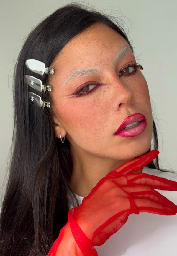 Femme aux longs cheveux noirs portant des barrettes argentées, un maquillage des yeux rose vif, un rouge à lèvres rouge éclatant et un gant rouge transparent près du visage.