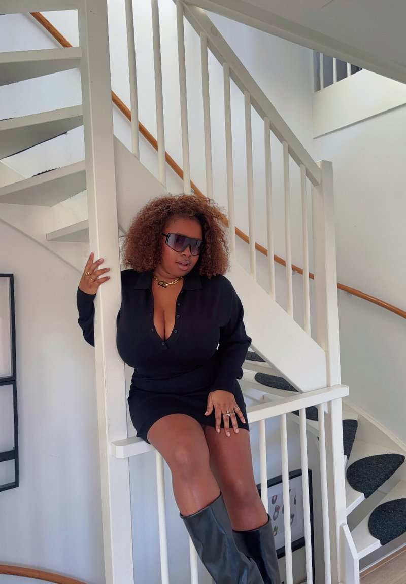 Robe noire à manches longues avec détails à boutons, assortie à des bottes noires montantes. Lunettes de soleil transparentes et bijoux en or complètent le look. Fond d'escalier blanc.