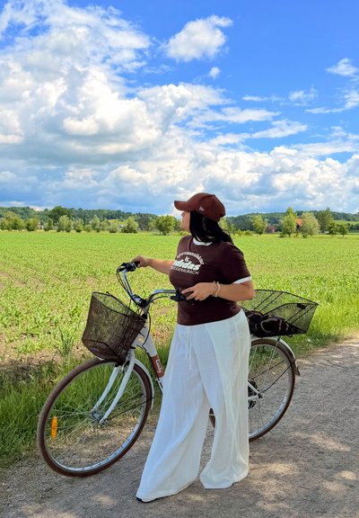 Mujer con camisa marrón y pantalones blancos de pie con una bicicleta en un camino junto a un campo verde bajo un cielo azul parcialmente nublado.