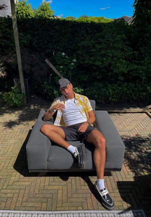 Sofá gris, persona sentada con las piernas extendidas, vestida con una camisa de cuadros amarillos sobre una camiseta blanca, pantalones cortos negros y zapatos negros con calcetines blancos.