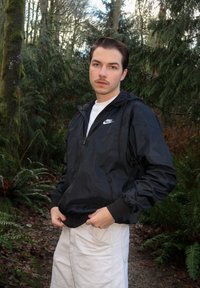 Coupe-vent en nylon noir avec une fermeture éclair à l'avant, une capuche ajustable et des poignets gris. Porté sur une chemise blanche, associé à un short en velours côtelé crème.