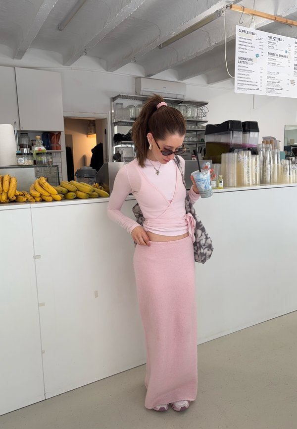 Moteris rožiniais drabužiais laiko gėrimą, stovi prie prekystalio su bananais ir permatomais puodeliais, modernioje kavinėje po meniu.