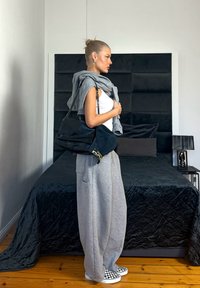 Femme en pantalon de survêtement gris et d ébardeur blanc se tient dans la chambre en tenant un grand sac à main noir et un sweat à capuche gris sur les épaules.