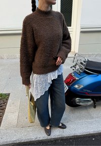 Rudas pūkuotas megztinis, uždėtas ant baltos nėriniuotos bluzonės, derinamas su plačiomis džinsinėmis kelnėmis ir rudais bateliais. Laiko žalią krepšį, šalia mėlyno motorolerio.