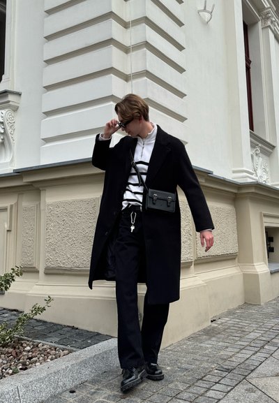 Abrigo negro sobre una sudadera blanca con cremallera y rayas negras, pantalones negros y botas negras. El accesorio incluye un pequeño bolso cruzado negro.