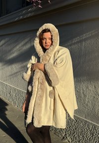 Roomkleurige poncho met imitatiebontvoering, voorzien van een capuchon, oversized mouwen en pomponaccents aan de voorkant. Zachte textuur, warme uitstraling.