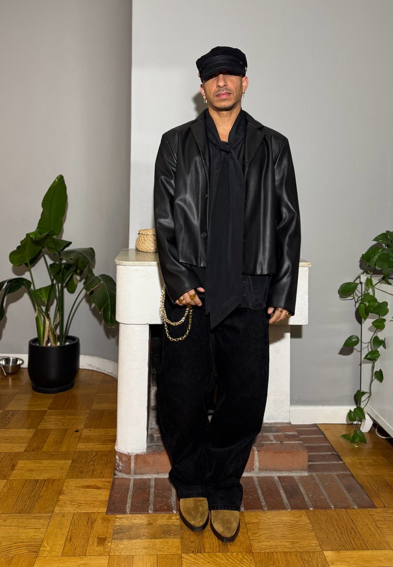 Giacca di pelle nera sopra una camicia nera con una sciarpa, abbinata a pantaloni larghi neri e scarpe marroni. Gli accessori includono una catena.