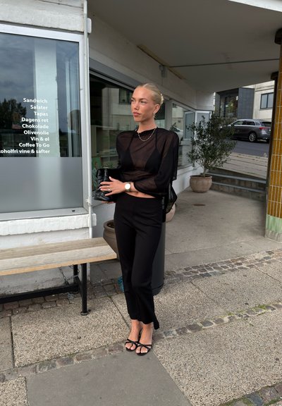 Top negro transparente con mangas largas, pantalones ajustados negros, sandalias negras con tiras, sosteniendo un bolso pequeño negro; diseño minimalista y esquema de color monocromático.