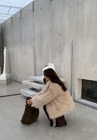 Cappotto in pelliccia sintetica beige con maniche larghe, pantaloni della tuta marroni e borsa verde oliva texture. Parete di cemento ed elementi scultorei sullo sfondo.