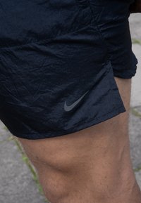Tumedasan sinine Nike lühikesed püksid, mis on valmistatud kerge kortsus kangast, sisaldavad hemil peent halli logo. Kujundus on lihtne ja funktsionaalne.