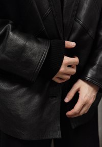 Veste en cuir noir à finition texturée, avec surpiqûres visibles, grands revers et coupe décontractée. Mains jointes devant.