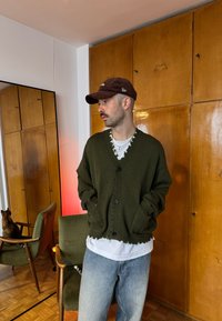 Homme en cardigan vert, chemise blanche et casquette debout à l'intérieur, les mains dans les poches, à côté d'une chaise verte et d'un chat tourné vers l'arrière.