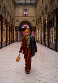 Completo in velluto a costine arancione con blazer oversize, camicia verde e cravatta fantasia. Porta una borsa marrone con accenti decorativi in una galleria.