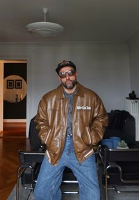 Brun fusk läder bomberjacka med vita ränder och text, i kombination med en grå skjorta och blå denimjeans. Runda solglasögon bar.