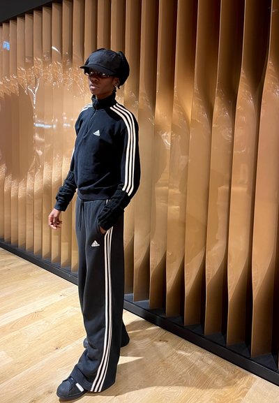 Chándal negro de Adidas con franjas blancas, que incluye una chaqueta con cremallera y pantalones de chándal. Gorro negro y zapatos negros texturizados. Suelo de madera lisa.