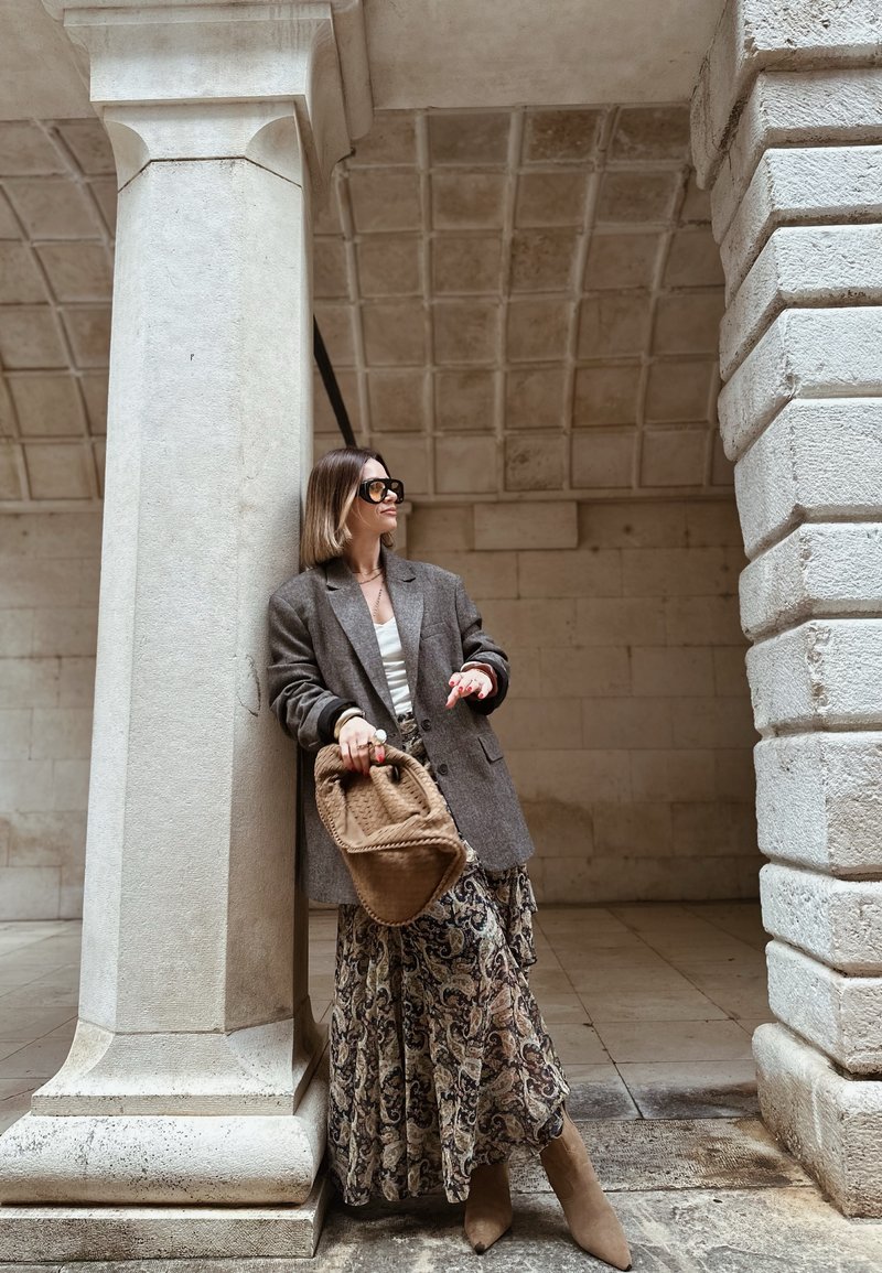 Femme portant un blazer gris surdimensionné, une jupe à motifs, des bottes beige et des lunettes de soleil, tenant un sac à main marron, adossée à une colonne en pierre.