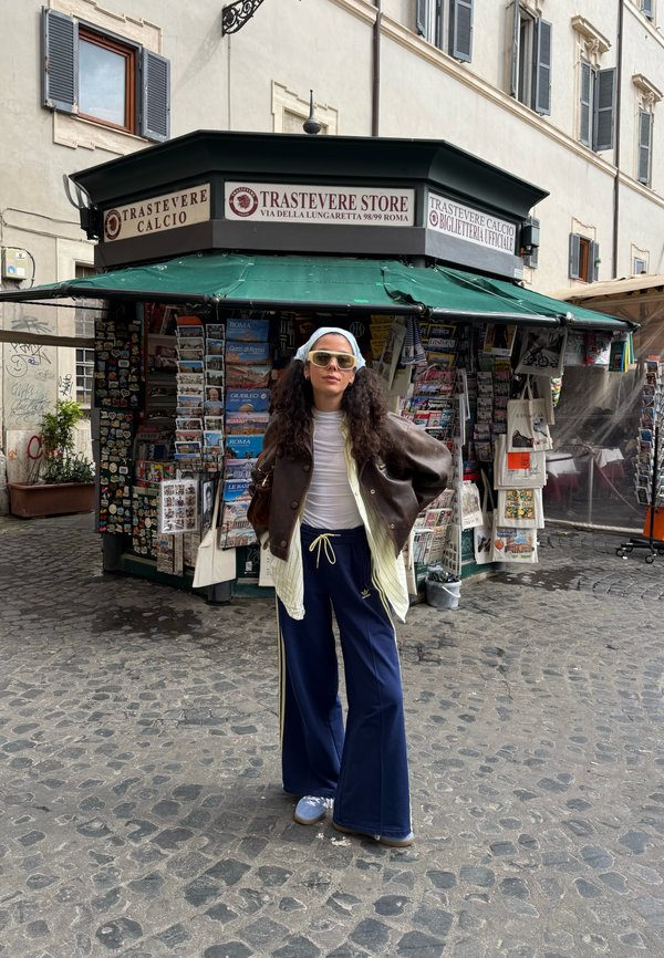 Una donna con una giacca di pelle marrone, una camicia bianca e pantaloni larghi blu navy si trova accanto a un chiosco di notizie con riviste e souvenir.