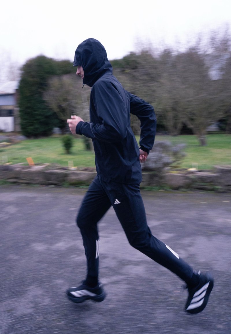 Person wearing black hooded jacket and pants running on paved path in cloudy, outdoor park setting.