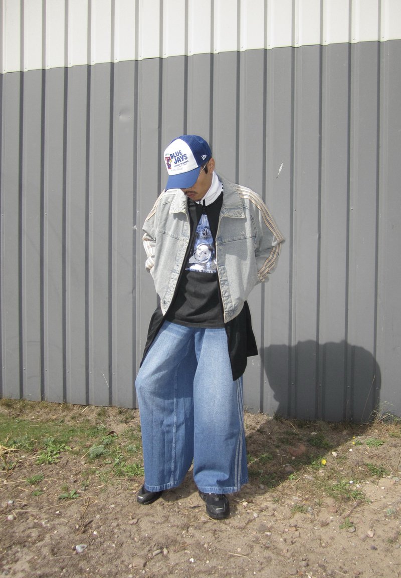 Veste en denim avec des manches rayées, t-shirt graphique noir en dessous, et jeans bleu à jambes larges. Casquette bleu et blanche, chaussures noires, debout à l'extérieur.