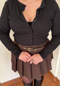 Camisa de manga larga negra con textura de canalé y botones, combinada con una falda marrón con pliegues y un cinturón marrón que presenta una hebilla y detalles dorados.