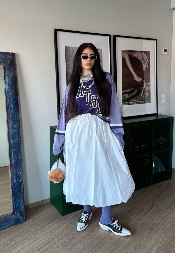 Femme aux longs cheveux foncés portant un haut de sport violet, une jupe blanche, des collants bleus, des baskets noires et des lunettes de soleil, tenant un porte-clés en peluche dans une pièce.