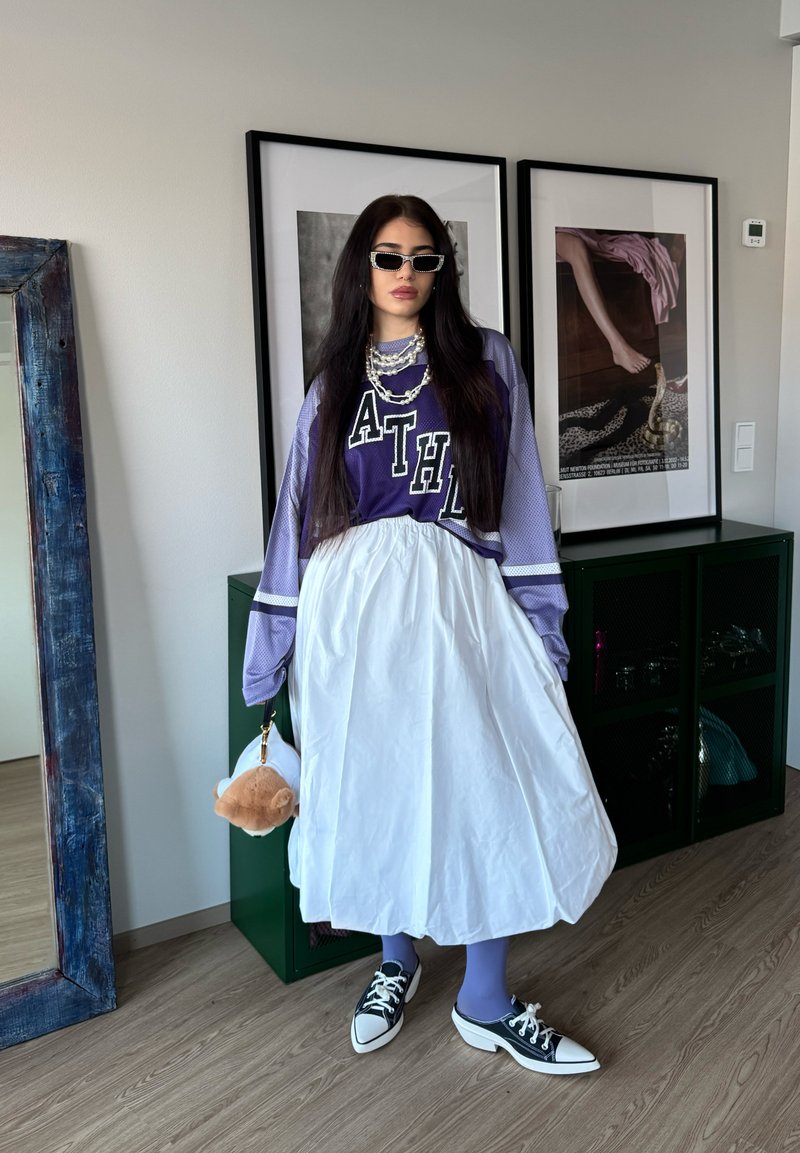 Femme aux longs cheveux foncés portant un haut de sport violet, une jupe blanche, des collants bleus, des baskets noires et des lunettes de soleil, tenant un porte-clés en peluche dans une pièce.