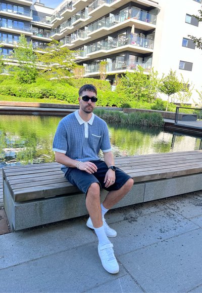 Camisa polo de manga corta gris con cuello y bolsillo blancos, pantalones cortos negros, zapatillas blancas, sentado en un banco de madera junto al agua con vegetación.
