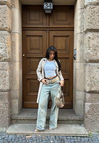 Beżowy trencz na biały krótki top, jasnoniebieskie szerokie jeansy oraz torebka w wzory. Stoi na kamiennych schodach przy drewnianych drzwiach.