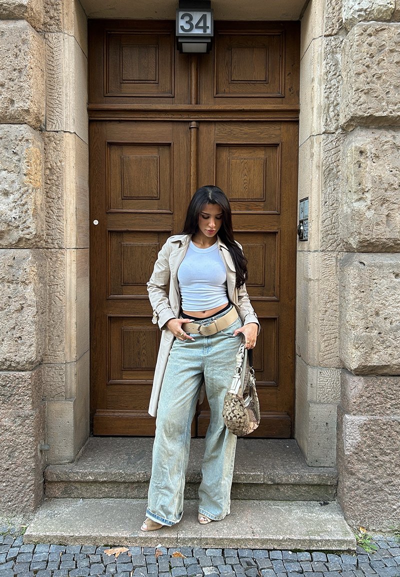Trench coat beige sopra un top corto bianco, jeans larghi azzurri e una borsa a motivo. In piedi su gradini di pietra di fronte a una porta di legno.