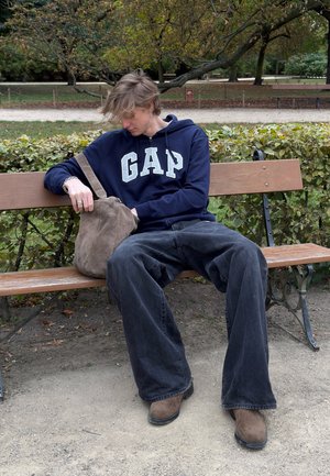 Modra kapuconasta majica z logotipom "GAP", ohlapni črni kavbojki, rjave semiš čevlje in rjava torba za ramena na klopi v parku.