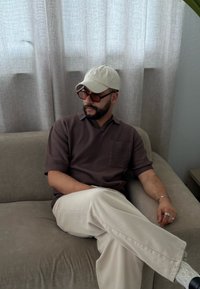 Brun kortermet polo skjorte med en teksturert lomme, lyse bukser og en hvit caps. Sitter på en grå sofa i et rom med nøytrale toner.
