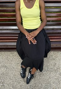 Żółty prążkowany top na ramiączkach, czarna plisowana spódnica, czarne trampki z białymi paskami oraz wyróżniający się pierścień i wzory na paznokciach. Siedząca na ławce.