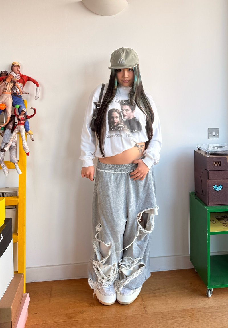 Jeune personne aux cheveux longs portant une casquette Adidas, un sweat-shirt graphique court, un pantalon de survêtement gris déchiré et des baskets blanches, debout à l'intérieur près d'étagères colorées.