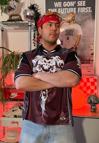 Jeune homme avec un bandana rouge, des lunettes de soleil sur la tête et les bras croisés, debout dans une pièce décorée avec des plantes, des chaussures et une affiche motivante.