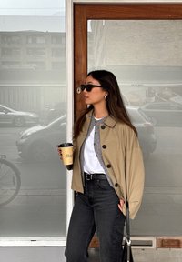 Tan oversized jakke med sorte knapper, grå cardigan under, hvid skjorte, sorte jeans og solbriller; holder en kaffekop.