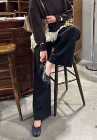 Zwarte wijde broek, donkerbruine trui en zwarte platte schoenen. Getextureerde bruine tas en zichtbare gelaagde kanten details. Houten kruk op de achtergrond.