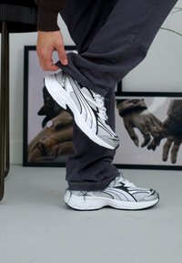Srebrni i bijeli sportski tenisice od mrežaste tkanine, s pojednostavljenim dizajnom i crnim akcentima. Nošene s sivim hlačama na minimalističkoj pozadini.