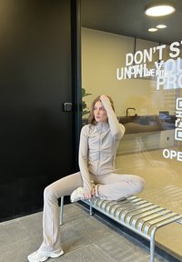 Survêtement beige ajusté avec veste zippée, tissu texturé et pantalons évasés. Assis sur un banc rayé argenté contre un mur noir.