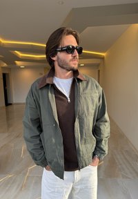 Chaqueta bomber verde oliva con cuello de pana marrón, cremallera frontal y dos bolsillos laterales. Llevada sobre un suéter marrón y pantalones blancos.