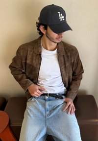 Brūns zamšādas mētelis ar apkakles kakla izgriezumu virs baltā T-krekla, kopā ar gaišiem džinsu biksēm. Apģērbam ir melns josta.