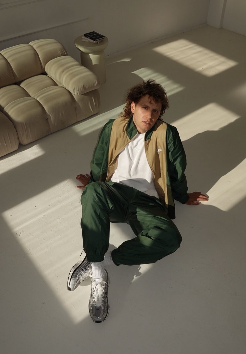 Sportliches Outfit mit einer grünen und beigen Jacke über einem weißen Hemd, kombiniert mit grünen Hosen und silbernen Sneakers. Der Hintergrund ist hell.