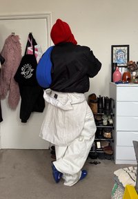 Personne vêtue de vêtements surdimensionnés superposés, avec une capuche rouge et des baskets bleues, se tenant dans une chambre où des chaussures et des vêtements sont visibles.