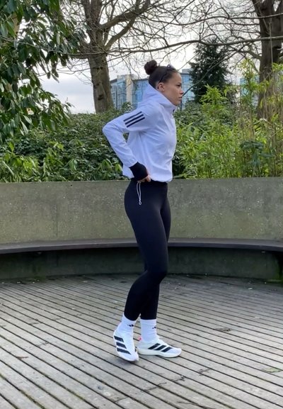 Atleta con una chaqueta blanca de cremallera con detalles negros, leggings negros y zapatillas blancas con rayas negras, de pie sobre una terraza de madera al aire libre.