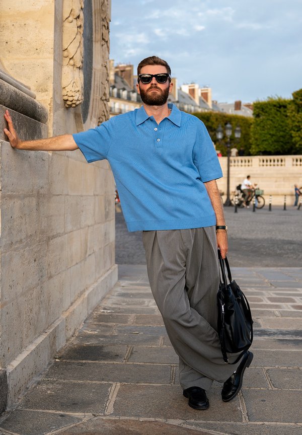 Man wearing sunglasses, blue shirt, and grey trousers leans on a stone wall, holding a black bag on a city street with buildings in the background.
