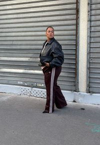 Casaco de couro oversized preto, camisa creme, calças de desporto bordô com riscas brancas e uma bolsa de mão tecida preta. Detalhe de fundo urbano.