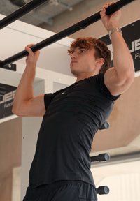 Svart atletisk skjorte og shorts; person som utfører en pull-up på en horisontal stang i et treningsstudio med grå bakgrunn.