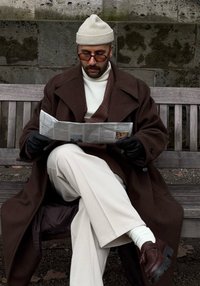 Καφέ μάλλινο παλτό, λευκή ζακέτα, καφέ και καρό ενδυμασία σε στρώσεις, ανοιχτά παντελόνια, μαύρα γάντια και σκούρα παπούτσια. Διαβάζει μια εφημερίδα.